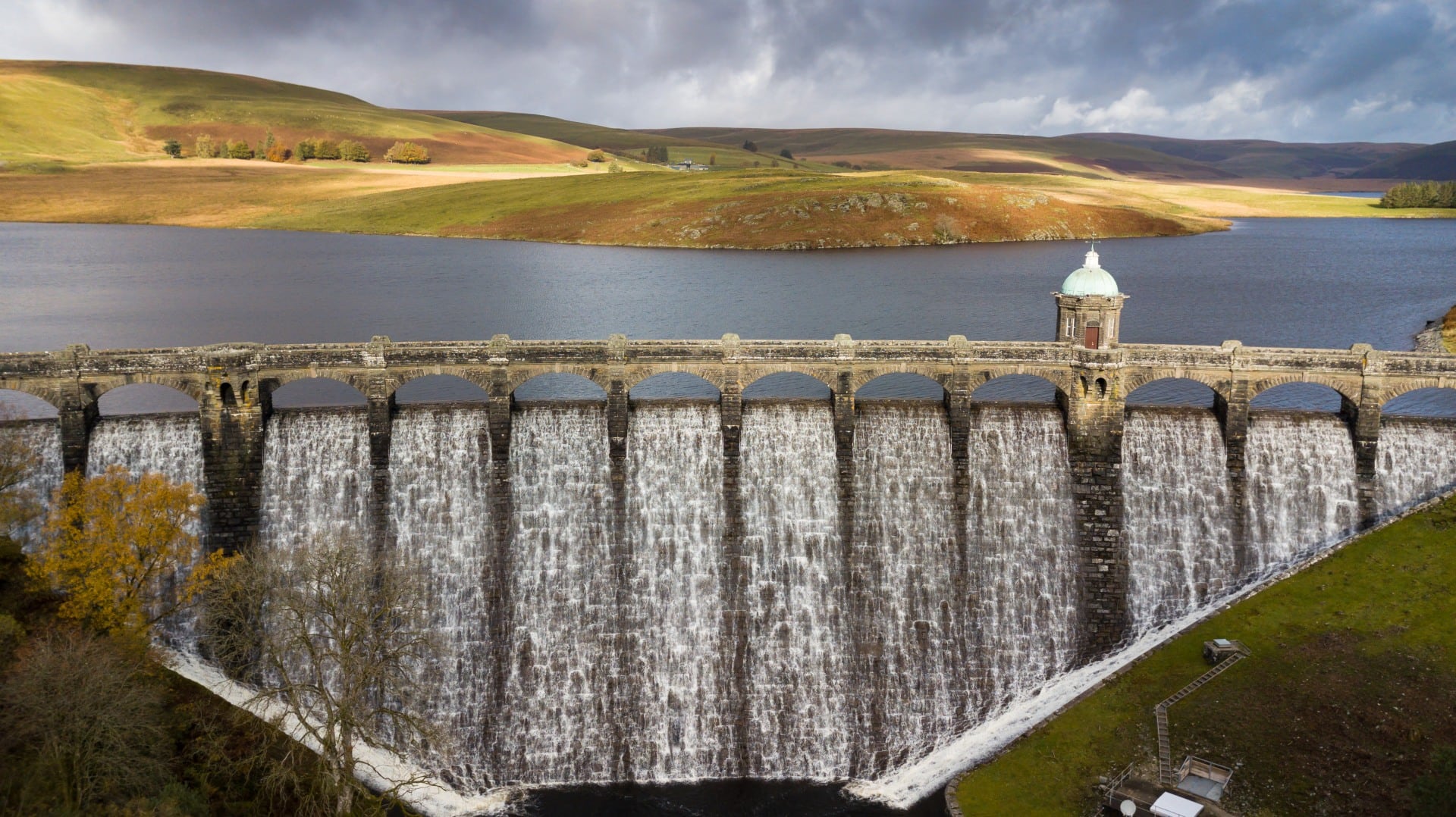 Water spills over Craig Goch dam beneath sunlit hills in the Elan Valley