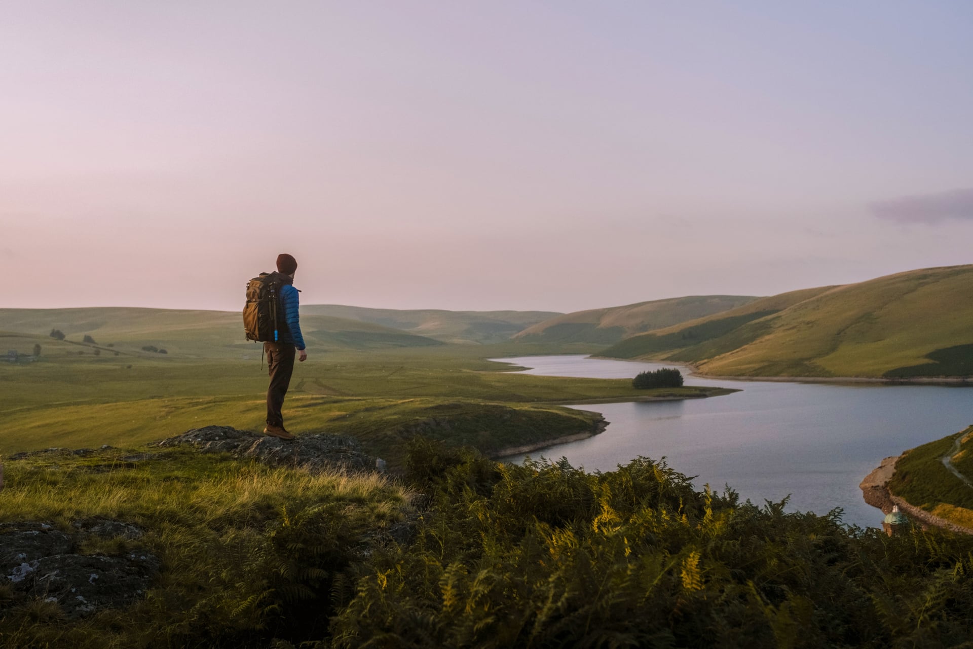 A walker pauses above a Welsh reservoir as soft evening light colours the hills